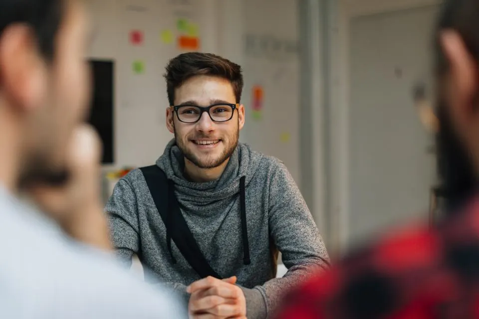 Um jovem de óculos conversando com 2 pessoas em um formato de entrevista de emprego